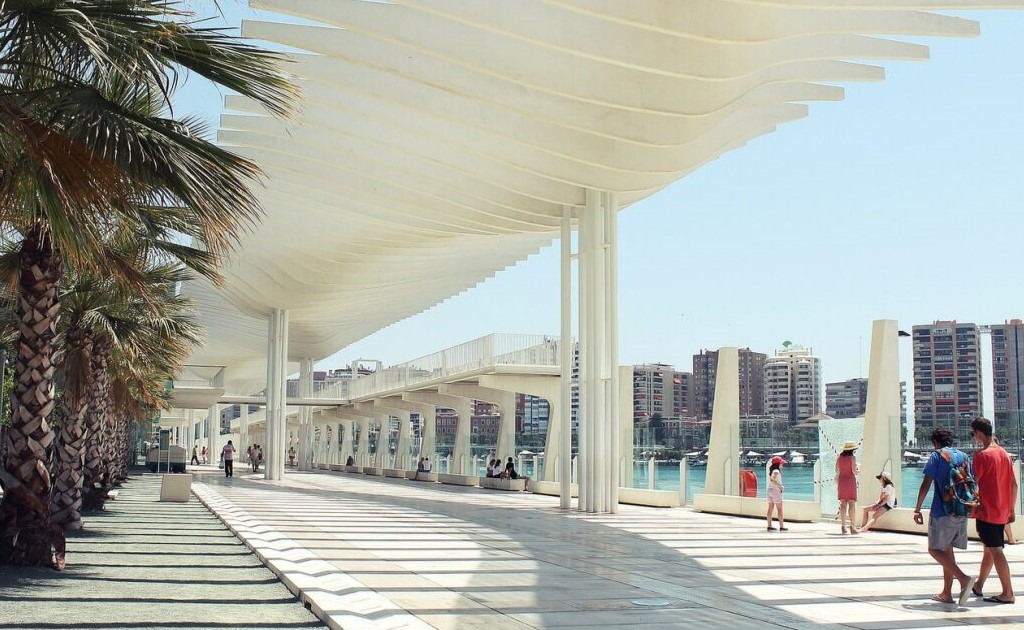 un paseo marítimo con personas transitándolo y palmeras a la izquierda, mientras tienes el mar y algunos apartamentos de fondo