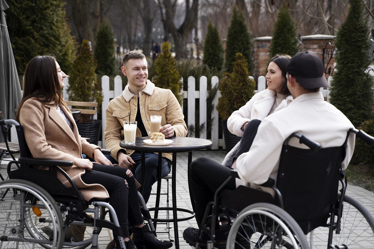 un grupo de personas, 2 de ellas en sillas de ruedas están en una terraza de un bar tomando unas bebidas, se encuentran felices.
