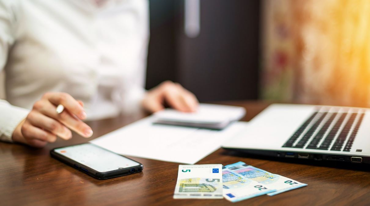 Mujer usando una calculadora encima de una mesa junto a su ordenador y unos billetes que tiene al lado.