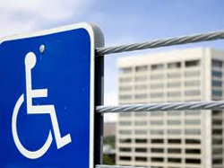 LIOUNDAU. SeñaL de discapacidad con una silueta de una persona en silla de ruedas conuna barrera metálica y de fondo un edificio de viviendas