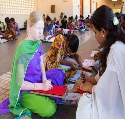 Grupo de mujeres, incluida una persona albina, realizan actividades educativas en el suelo, usando herramientas y cuadernos. Están en un espacio amplio, con luz natural y ambiente colaborativo.