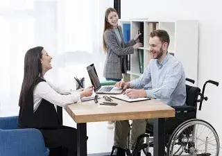 Una persona con discapacidad y en silla de ruedas, trabajando con dos de sus compañeras en la oficina (Fuente: Servimedia)